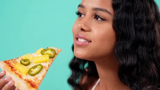 Joyful moment of a woman savoring a pizza slice with pineapple and jalapenos against a turquoise background