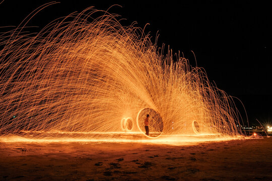 Amazing steel wool fire show performance on the beach at night. Long exposure photography capturing glowing orange sparks and spinning light circles near the sea.