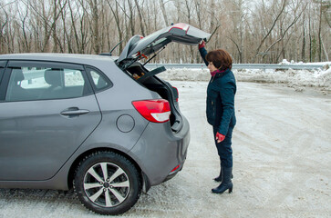 Senior woman driver opens the trunk of car on snowy winter road, preparing to pick up luggage or groceries in city street parking lot.