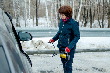 Senior woman driver standing on snowy roadside with portable air compressor, preparing to inflate car tire in city street parking lot.
