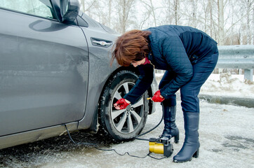 Elderly woman beside gray car on icy road, crouching to check and pump tire pressure with compact compressor during snowy trip, highlighting winter breakdown service and careful driver behavior.