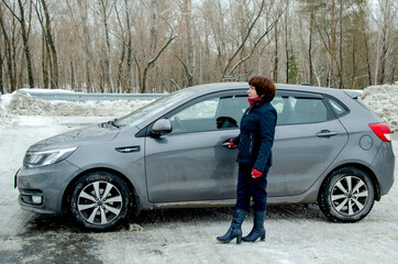 Attractive senior woman driver standing beside gray car on snowy roadside near metal guardrail, looking ahead in cold winter landscape.