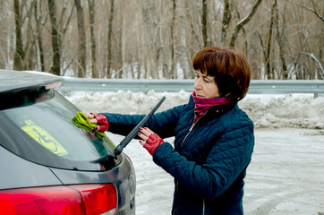 Senior woman cleaning rear car window with green cloth and raised wiper on snowy outdoor parking lot, focusing on safe visibility, winter vehicle maintenance
