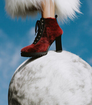 A red high heel boot with plush furry texture stands on a fluffy white and grey orb beneath a clear blue sky. Luxe winter fashion aesthetic with dramatic elevation.
