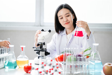 This image shows a female scientist in a laboratory wearing a white lab coat. She is examining a flask containing a red liquid while sitting next to a microscope. On the table, there are various labor