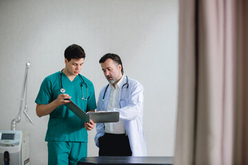 two male doctors in a hospital room reviewing medical documents together. One doctor is wearing green scrubs with a stethoscope around his neck, while the other is in a white coat, also with a stethos