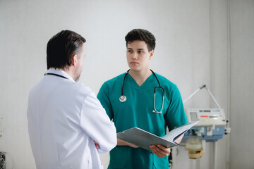 two male doctors in a hospital room reviewing medical documents together. One doctor is wearing green scrubs with a stethoscope around his neck, while the other is in a white coat, also with a stethos