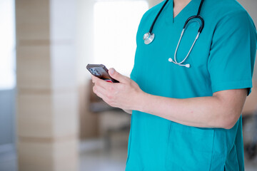 Male doctor wearing lab coat holding cellphone beside windows in hospital