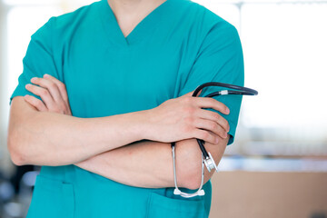 Close-up of unrecognizable doctor holding stethoscope while standing with arms crossed at medical clinic.