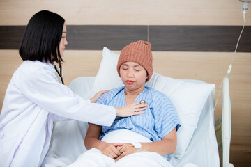 A compassionate doctor examines a female patient wearing a knitted hat in a hospital bed, checking her heartbeat with a stethoscope, showing care and medical attention during treatment.