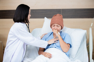 A compassionate doctor examines a female patient wearing a knitted hat in a hospital bed, checking her heartbeat with a stethoscope, showing care and medical attention during treatment.