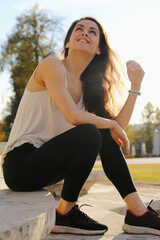 Happy beauty face woman with long hair looking in camera sitting in casual clothes on stairs outdoor summer bright sun background. Closeup fun relaxing person street portrait