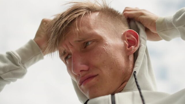 low shot white male tightening hood, cloudy sky backdrop, upward camera angle, determined expression, drawstrings pulled tight, portrait of preparation and resolve, outdoor ritual before run,