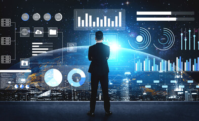 A businessman stands in front of a futuristic digital interface displaying graphs, charts, and data visualization elements against an urban skyline at night. Gantry