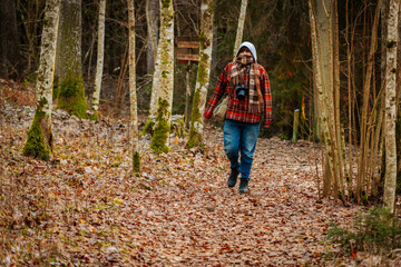 Fototapeta premium Individual walking through a serene forest path, surrounded by autumn foliage and tall trees, wearing a plaid jacket and capturing nature with a camera, showcasing outdoor exploration