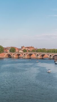 Garonne River and Pont Neuf timelapse with Port de la Daurade in downtown Toulouse, France. This Renaissance arch bridge reflects in the water under a blue sky with clouds. Waterfront with green trees