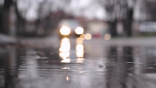 Headlights bokeh and lights reflections of street traffic in puddle while raindrops falling