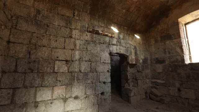 Atmospheric interior of an ancient stone ruin, featuring weathered block walls, a vaulted ceiling, and small windows allowing light rays to pierce the historic gloom. 