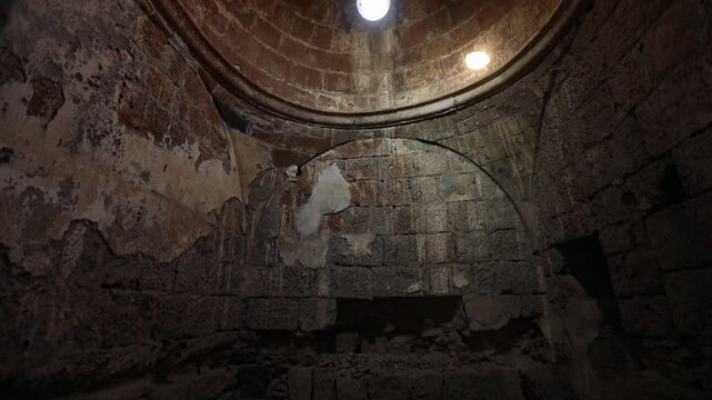 Atmospheric interior of an ancient stone ruin, featuring weathered block walls, a vaulted ceiling, and small windows allowing light rays to pierce the historic gloom. 