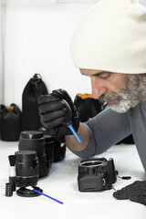 A man in a white beanie and black gloves uses a rubber air blower to clean a camera sensor. Multiple lenses and cleaning tools are neatly arranged on the white desk in front of him.