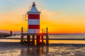 Vecchio Faro lighthouse in Lignano Sabbiadoro at sunrise, red and white beacon above shallow sea with reflections, iconic Adriatic coast landmark in Italy, calm travel and tourism concept.