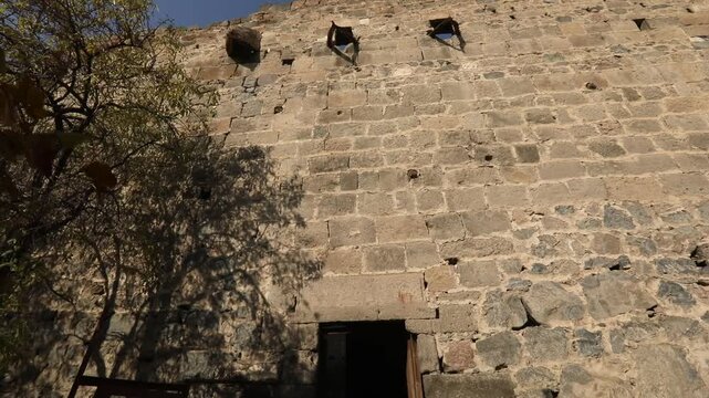 This video captures the timeless beauty of ancient stone ruins, likely a historical church or monastery, under a clear blue sky. Bare tree branches with lingering autumn leaves frame the weathered wal