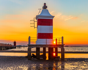 Vertical view of Vecchio Faro lighthouse at sunrise in Lignano Sabbiadoro, red and white beacon above shallow sea with reflections, iconic Adriatic coast landmark in Italy, calm travel concept.
