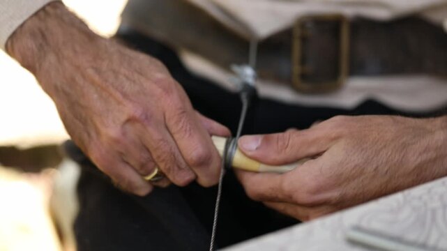 Close-up of male hands wrapping black thread around a wooden and brass tool handle. A craftsman carefully repairs a small instrument part with manual skill and precision.

