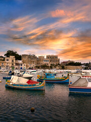 View of Birzebbuga's small harbor with numerous moored boats at sunset ; rows of houses appear in the background.