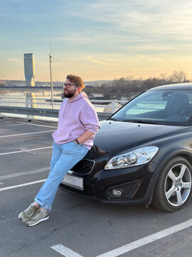 Man leaning on car near river at sunset. Urban lifestyle, freedom, modern masculinity, and reflective pause in city environment