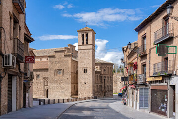 Obraz premium The Mudéjar tower of Santiago del Arrabal stands on the left, leading toward the Puerta de Bisagra gate in Toledo. Brick buildings and shops line the right side