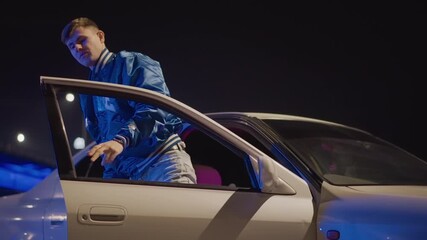 young caucasian man stepping out of car under neon bridge lights, white sedan parked on roadside, blue varsity jacket, hands on hips, moody cinematic glow and solitary street vibe