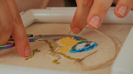A close-up of hands working on embroidery, using rainbow scissors to trim threads on a fabric canvas featuring colorful stitched patterns. The focus is on the delicate craftsmanship