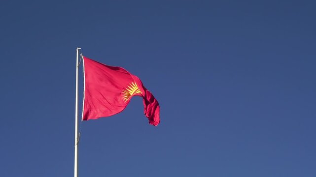 Red banner featuring a central golden sun with forty rays and a tunduk pattern, representing unity, heritage, and the nomadic culture of Kyrgyzstan, fluttering prominently in open daylight.