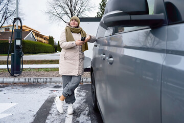 Calm independence is visible as a middle aged woman waits beside her electric car while checking her phone, turning charging time into a quiet personal pause.