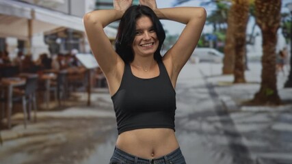 Woman with bare midriff wearing black tank top smiling while making bunny ears gesture on sunlit street; fun.