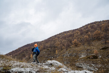 Naklejka premium young hiker standing on ridge against moody sky, lone figure in bright jacket surveying distant wooded hillside, sense of independence and fresh air, autumn tones and textured rock underfoot
