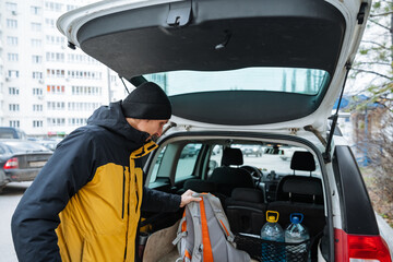 caucasian man leaning into trunk checking supplies and backpack, adjusting bottles and straps, urban residential backdrop with autumn foliage, purposeful preparation for outdoor task