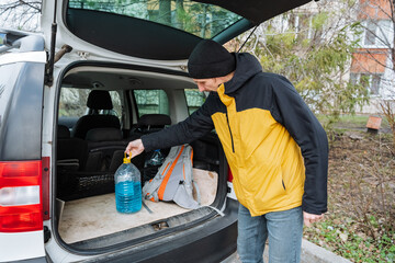 organizing supplies in urban vehicle environment, man carefully loads water container into vehicle trunk, urban setting with man preparing emergency water stock in vehicle