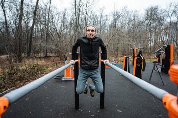 man performing parallel bar dip in outdoor gym, controlled descent and explosive press, casual coldweather layers, bare trees and orange equipment frame urban strength routine