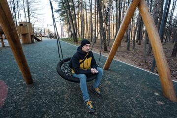 gentleman relaxes on outdoor swing seat, stylish male resting quietly on swing amidst park scenery, seated man in beanie and jacket enjoying tranquil park environment in natural light