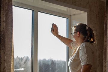 A woman washes a window in a house with a microfiber cloth. The woman is doing housework while bright sunlight streams into the room. The woman is washing the window.