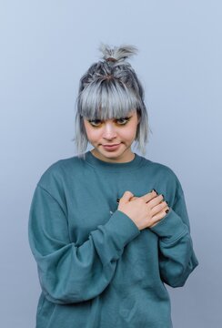 Person with gray hair and a green sweater stands with hands over heart against a light blue background while looking slightly down