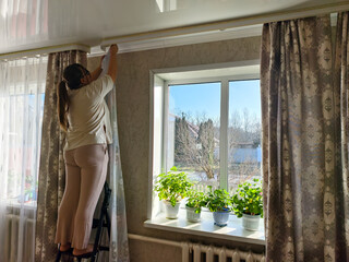 A woman stands on a stepladder, busily hanging curtains on a rod in her sunlit living room. While doing her chores, she completes her home decor with new window treatments.