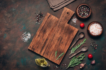 An empty wooden cutting board with various spices, herbs, and seasonings on a dark rusty grunge background. A culinary backdrop with cooking ingredients. Top view, flat lay, copy space.