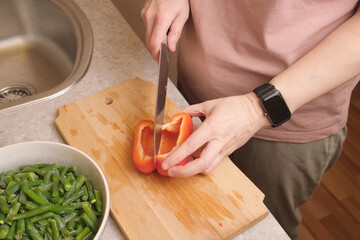 Woman chopping red sweet pepper on a wooden cutting board beside a bowl of frozen green beans in a cozy kitchen, preparing vegetables for a hot homemade dish for recipe blogs and family dinner ideas