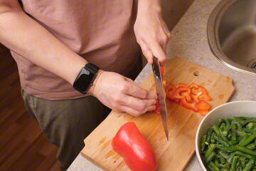 Woman chopping red sweet pepper on a wooden cutting board beside a bowl of frozen green beans in a cozy kitchen, preparing vegetables for a hot homemade dish for recipe blogs and family dinner ideas