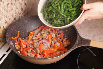 Home cook pours frozen green beans from a bowl into a sizzling wok with browned meat, red pepper, and onion, creating a colorful balanced stir fry for healthy dinner, recipe videos