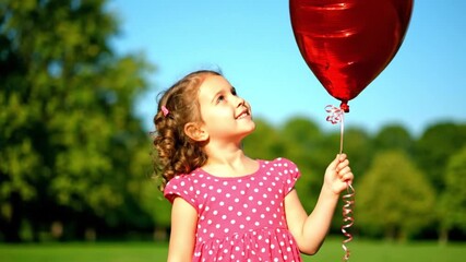 Smiling girl in pink dress holding red heart balloon on a sunny day in nature