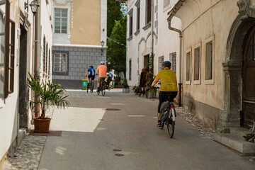 Cyclists ride through a charming street in Radovljica's old town, flanked by historic medieval buildings with colorful facades. The cobblestone road curves past authentic stone architecture under a